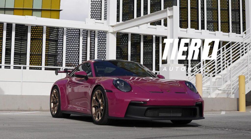 A magenta Porsche sports car with bronze rims parked in front of a modern white and yellow building structure, featuring the logo 'TIER 1 AUTOHAUS' prominently displayed in the foreground.