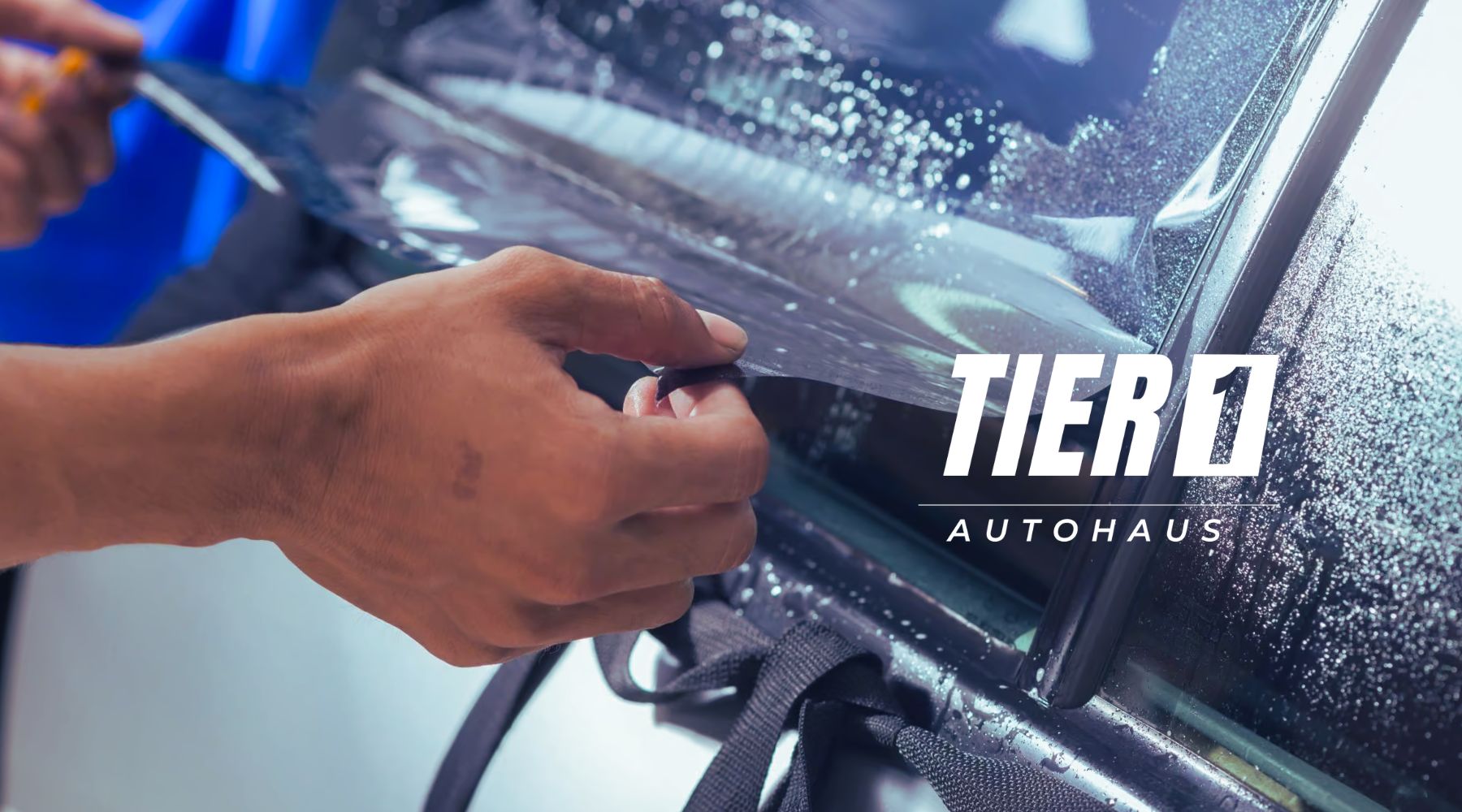 DIY Window Tinting - a person's hands applying a sheet of window tint film to a car window with precision. The logo "TIER 1 AUTOHAUS" is visible on the right side of the image, indicating the brand or service associated with the tinting process. The window is wet, likely to assist in the application of the tint film