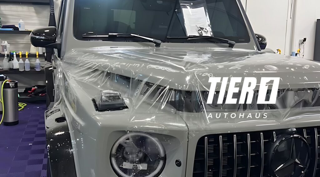 A gray Mercedes-Benz G-Class with paint protection film partially applied on the front, with the text "TIER1 AUTOHAUS" overlaying the image, emphasizing the brand involved in the installation.